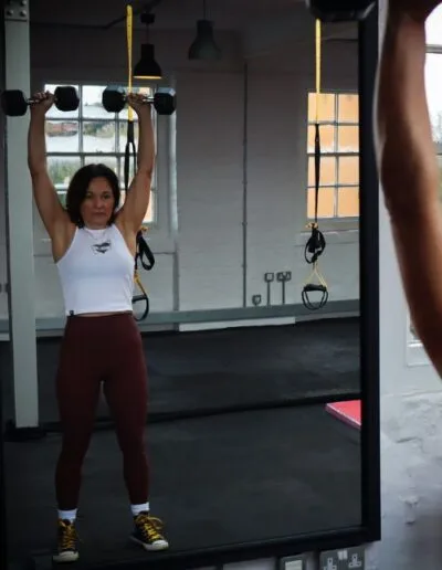 Woman in her 50s wearing brown leggings and white top lifting weights and looking at herself in mirror at womens gym in Derby